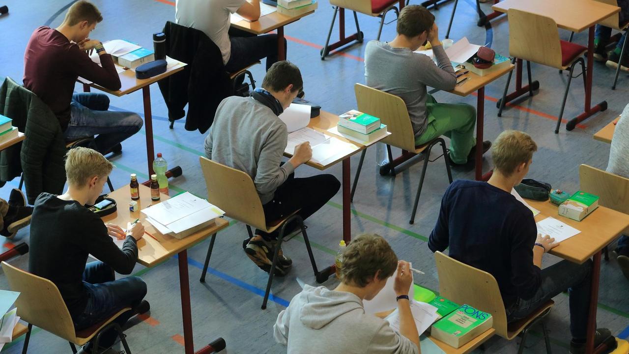 Schülerinnen und Schüler sitzen in einer Turnhalle an Einzeltischen und machen Abiturprüfungen. Vor ihnen liegen Englisch-Wörterbücher. Schülerinnen und Schüler sitzen in einer Turnhalle an Einzeltischen und machen Abiturprüfungen. Vor ihnen liegen Englisch-Wörterbücher.