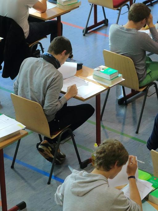 Schülerinnen und Schüler sitzen in einer Turnhalle an Einzeltischen und machen Abiturprüfungen. Vor ihnen liegen Englisch-Wörterbücher.