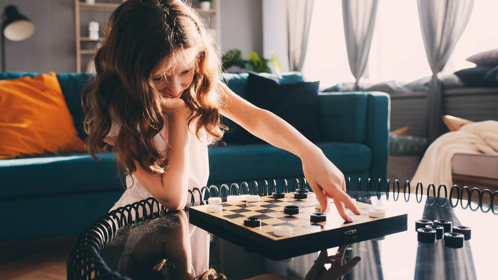 Ein Mädchen verschiebt mit der linken Hand einen Spielstein auf einem Brettspiel, ihr Kinn hat sie auf die rechte Hand gestützt.