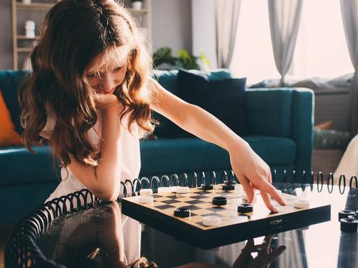 Ein Mädchen verschiebt mit der linken Hand einen Spielstein auf einem Brettspiel, ihr Kinn hat sie auf die rechte Hand gestützt.