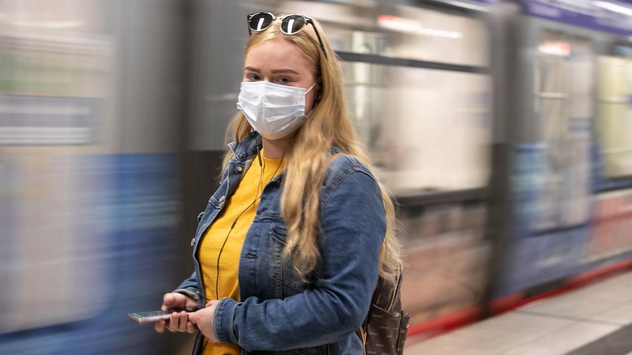 Eine Frau mit einem Mund-Nase-Schutz steht vor einer einfahrenden U-Bahn. Eine Frau mit einem Mund-Nase-Schutz steht vor einer einfahrenden U-Bahn.