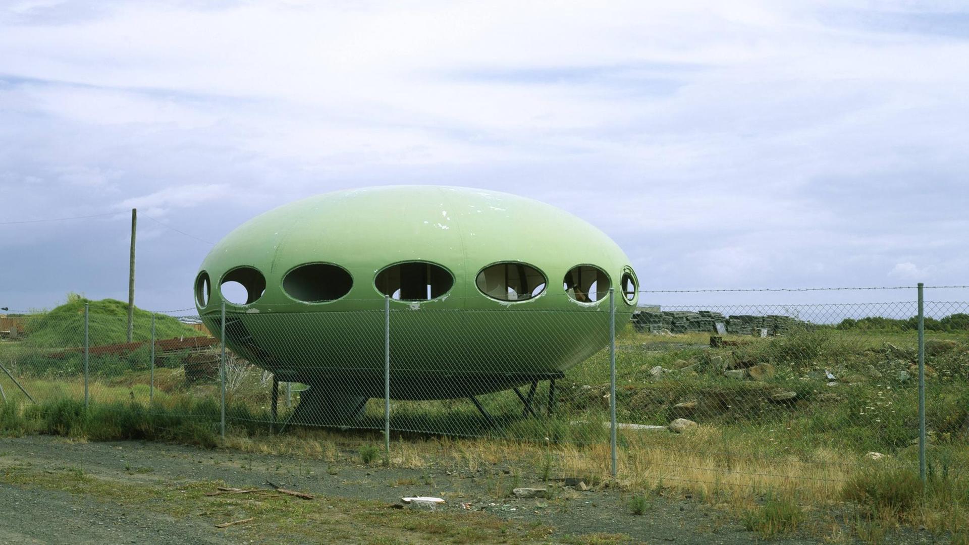 Ein grünes Futuro House steht hinter einem Zaun in einer industriellen Brachlandschaft. Ein grünes Futuro House steht hinter einem Zaun in einer industriellen Brachlandschaft.
