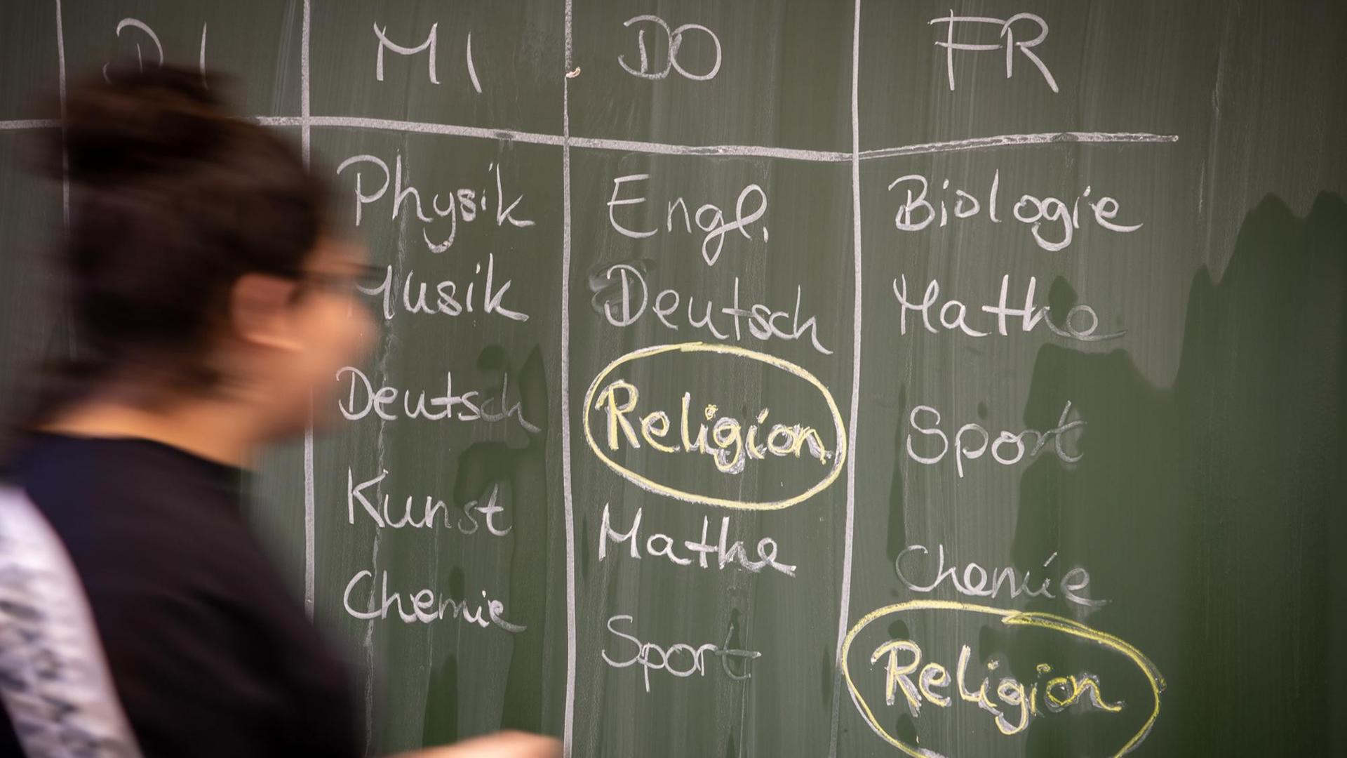 Eine Schülerin steht an einer Tafel, auf der ein Stundenplan mit Religionsunterricht geschrieben ist. Das Fach ist umkreist. Eine Schülerin steht an einer Tafel, auf der ein Stundenplan mit Religionsunterricht geschrieben ist. Das Fach ist umkreist.