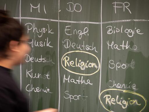 Eine Schülerin steht an einer Tafel, auf der ein Stundenplan mit Religionsunterricht geschrieben ist. Das Fach ist umkreist.