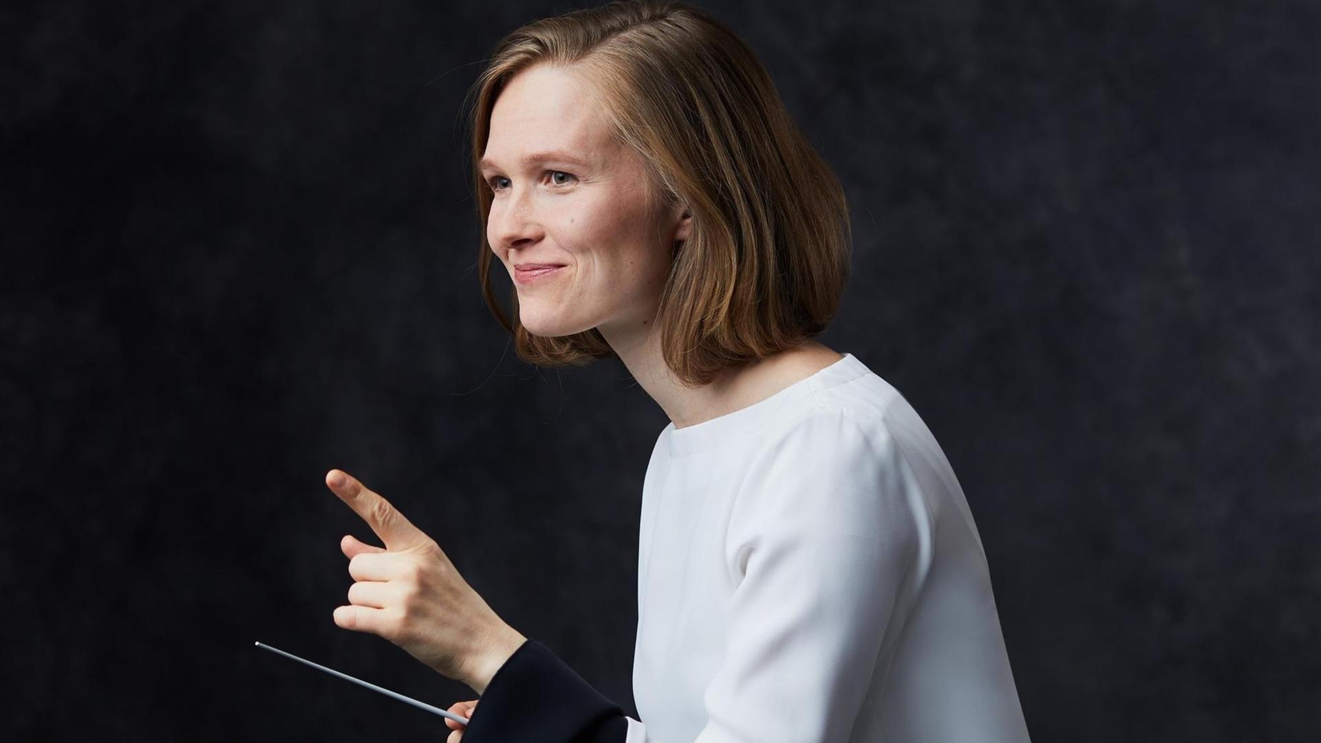 Eine junge Frau blickt freundlich und hebt den linken Zeigefinger. In der anderen Hand hält sie einen Dirigierstab. Eine junge Frau blickt freundlich und hebt den linken Zeigefinger. In der anderen Hand hält sie einen Dirigierstab.