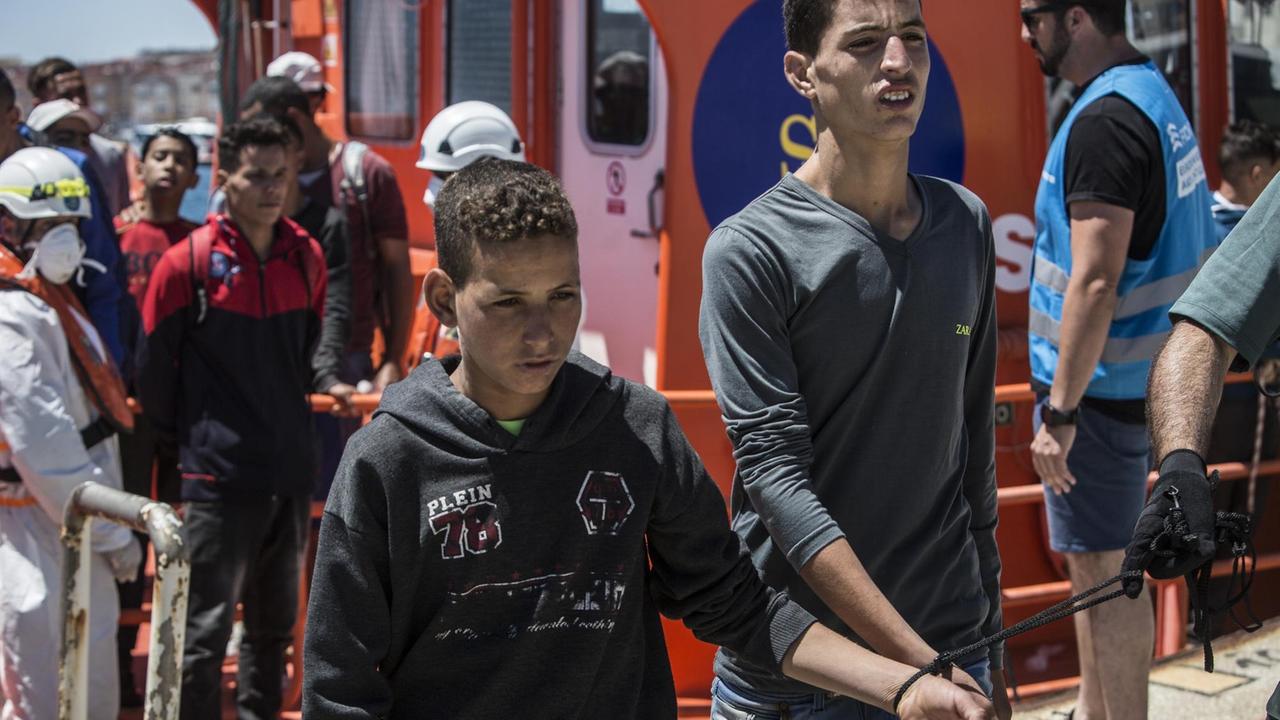 29.06.2018, Spanien, Tarifa: Zwei junge Migranten aus Nordafrika kommen nach ihrer Rettung in der Straße von Gibraltar im Hafen von Tarifa an und werden von einem Polizisten begleitet. Ihre Hände wurden zuvor zusammengebunden. Die EU-Staaten haben sich bei ihrem Gipfel in Brüssel darauf geeinigt, in der EU geschlossene Aufnahmelager für gerettete Bootsflüchtlinge einzurichten. Foto: Javier Fergo/dpa | Verwendung weltweit