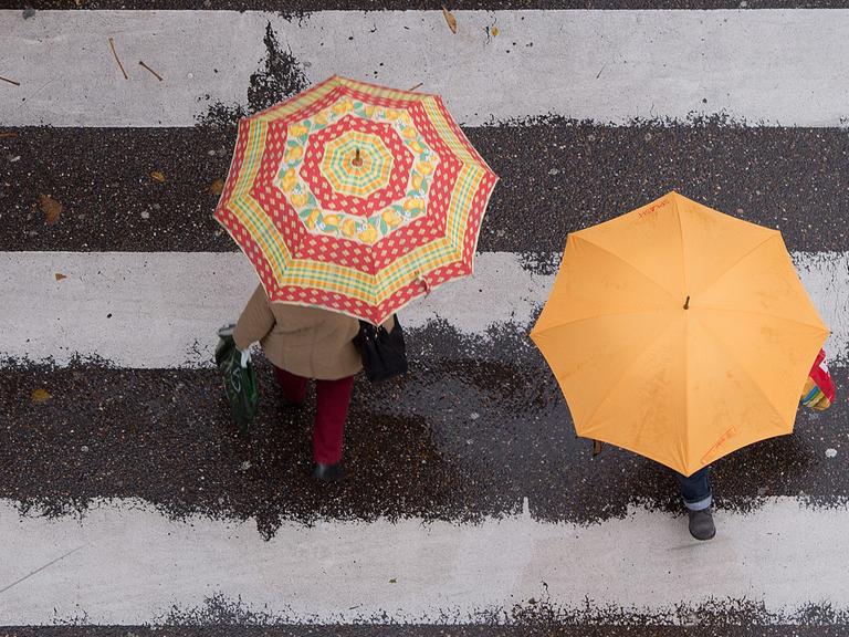 Zwei Passanten mit farbigen Regenschirmen gehen in Stuttgart über einen Zebrastreifen.