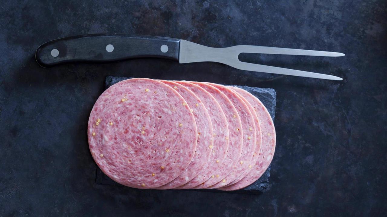 Rosa gesprenkelte Wurstscheiben liegen mit einer Wurstgabel auf einer Schieferplatte. Rosa gesprenkelte Wurstscheiben liegen mit einer Wurstgabel auf einer Schieferplatte.