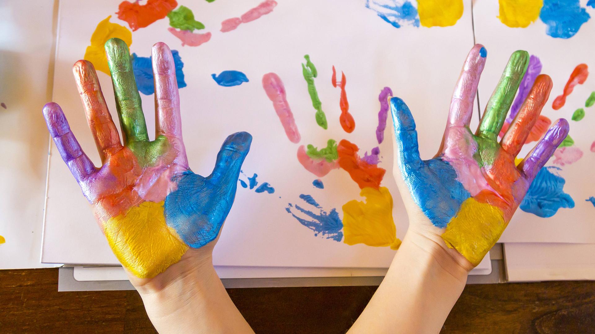 Zwei Hände mit sechs Farben bemalt werden in die Kamera gehalten, im Hintergrund Papier mit den Abdrücken der Hände.