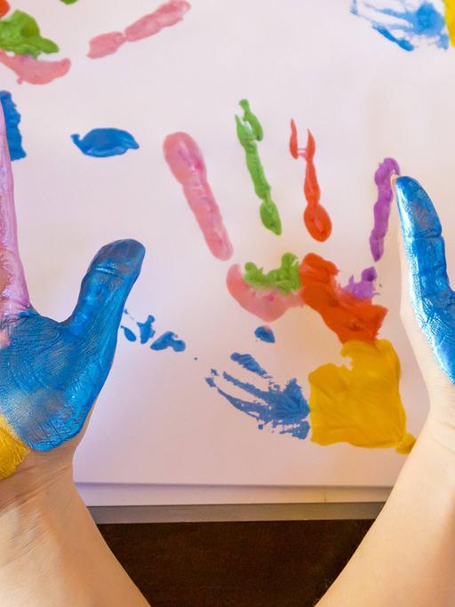 Zwei Hände mit sechs Farben bemalt werden in die Kamera gehalten, im Hintergrund Papier mit den Abdrücken der Hände.