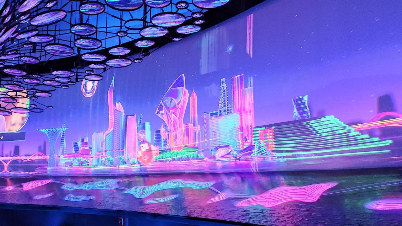 Ein futuristisch anmutender Raum in den Farben Neonpink-, blau und lila. An eine Leinwand ist eine Stadt projiziert, wie sie in der Zukunft aussehen könnte. Ein futuristisch anmutender Raum in den Farben Neonpink-, blau und lila. An eine Leinwand ist eine Stadt projiziert, wie sie in der Zukunft aussehen könnte.