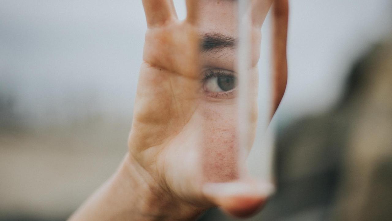 Eine Hand hält einen Spiegel, in dem der Auschnitt eines Gesichts mit einem Auge zu sehen ist.