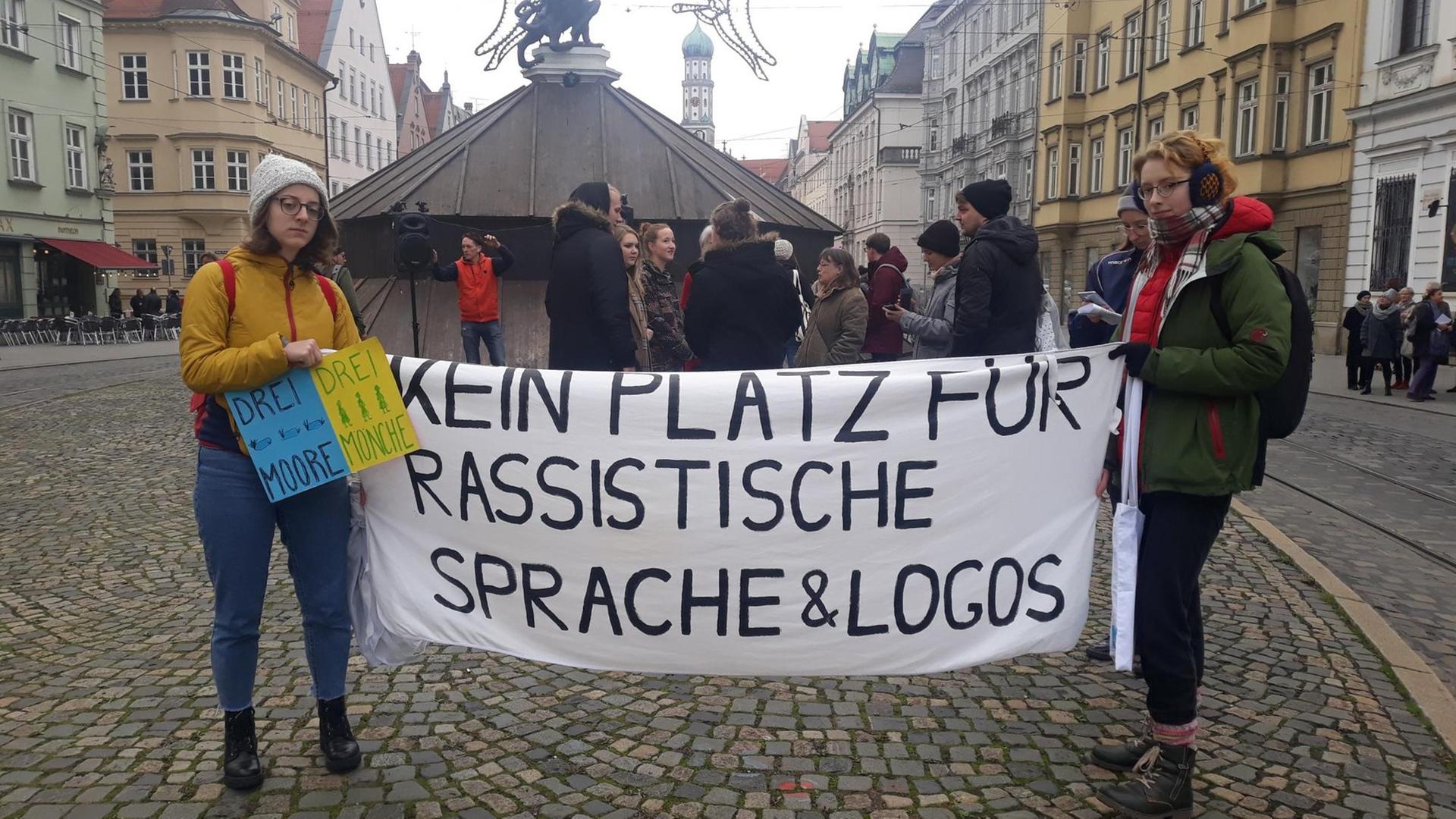 Zwei Menschen halten in Augsburg bei einer Kundgebung zur Umbenennung des Hotels "Drei Mohren" ein Banner mit der Aufschrift "Kein Platz für rassistische Sprache & Logos" zwischen sich, im Hintergrund weitere Personen und ein Brunnen. Zwei Menschen halten in Augsburg bei einer Kundgebung zur Umbenennung des Hotels "Drei Mohren" ein Banner mit der Aufschrift "Kein Platz für rassistische Sprache & Logos" zwischen sich, im Hintergrund weitere Personen und ein Brunnen.