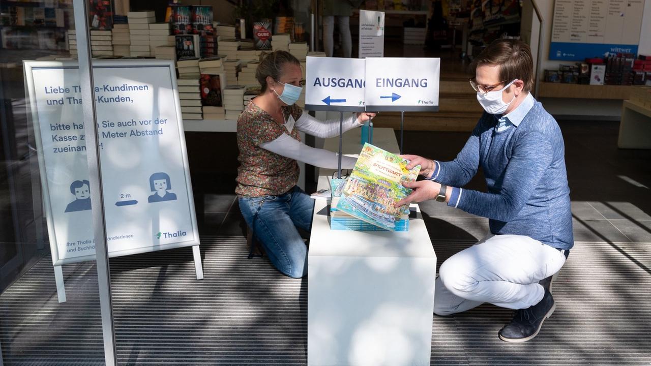 Ein Mann und eine Frau tragen Schutzmasken aus Stoff vor dem Gesicht und platzieren Bücher auf dem Podest vor dem Eingang einer Buchhandlung. Ein Schild hinter ihnen weist Kunden darauf hin, dass sie beim Betreten des Geschäfts voneinander Abstand halten sollen. Ein Mann und eine Frau tragen Schutzmasken aus Stoff vor dem Gesicht und platzieren Bücher auf dem Podest vor dem Eingang einer Buchhandlung. Ein Schild hinter ihnen weist Kunden darauf hin, dass sie beim Betreten des Geschäfts voneinander Abstand halten sollen.