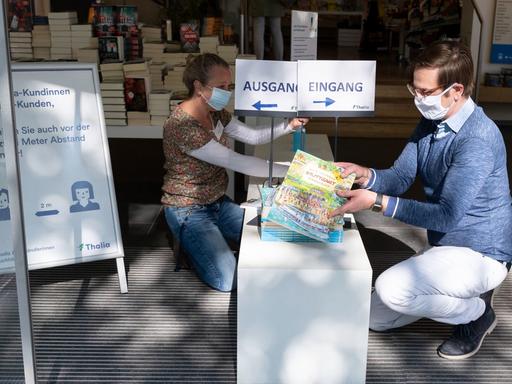 Ein Mann und eine Frau tragen Schutzmasken aus Stoff vor dem Gesicht und platzieren Bücher auf dem Podest vor dem Eingang einer Buchhandlung. Ein Schild hinter ihnen weist Kunden darauf hin, dass sie beim Betreten des Geschäfts voneinander Abstand halten sollen.