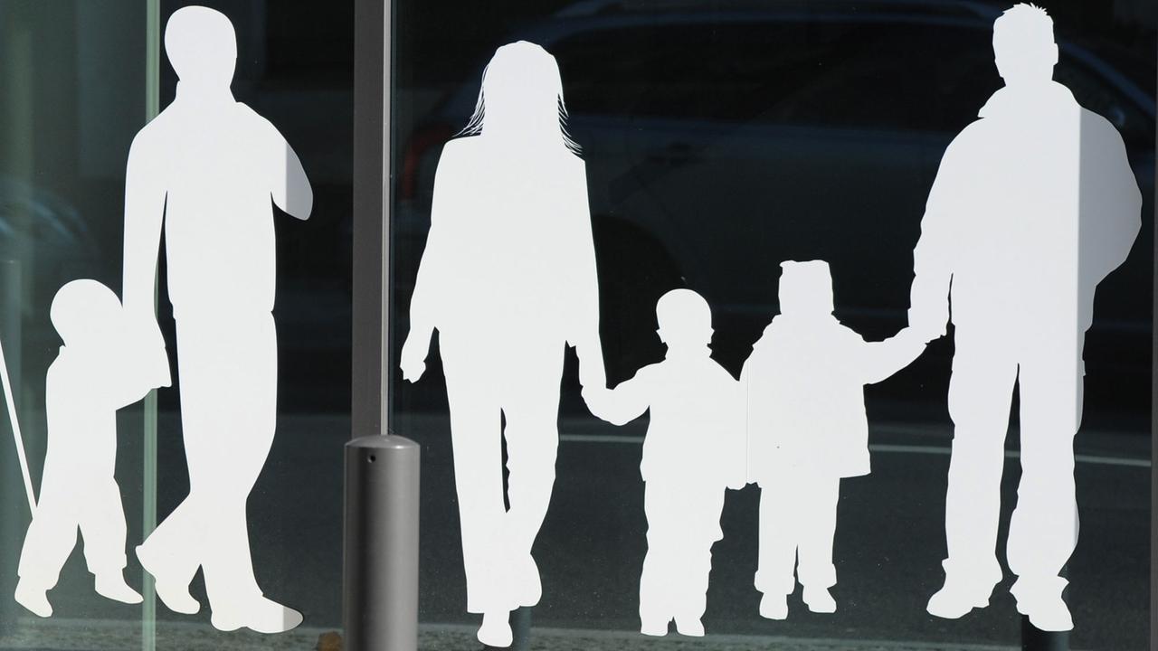 Weiße Figuren von einem Vater mit Kind und einer Familie mit Kindern kleben auf einer Glasscheibe. 