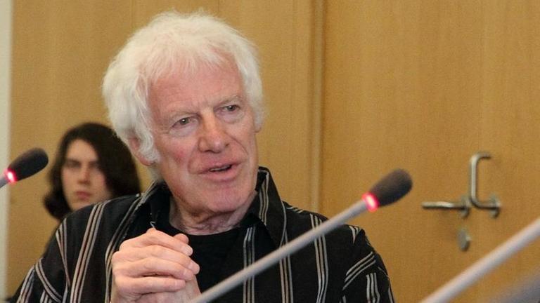 Ein Mann mit weißen Haaren spricht mit verschränkten Händen hinter einem Mikrophon Ein Mann mit weißen Haaren spricht mit verschränkten Händen hinter einem Mikrophon