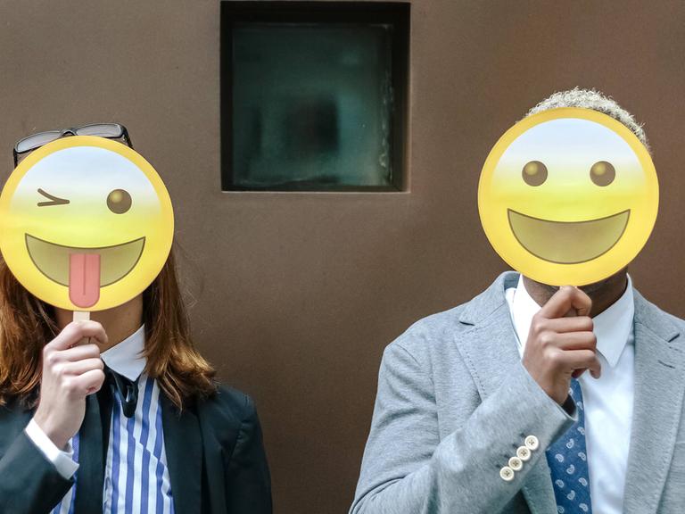 Eine Frau und ein Mann verdecken ihre Gesicher mit Emoji-Masken