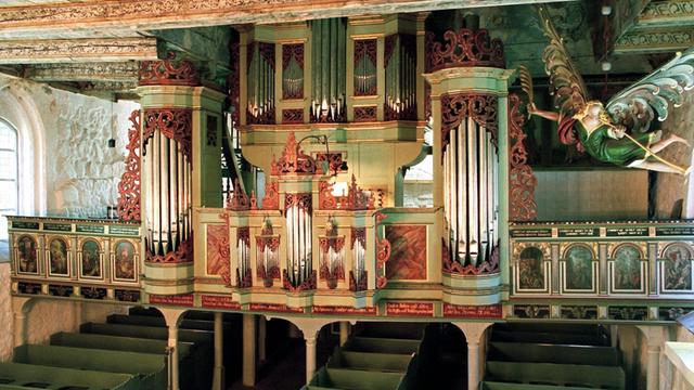 Das historische Instrument ist von grün-, rot-, goldfarbenem Holz umgeben, links und rechts befindet sich jeweils eine Empore, auf deren Aussenseiten gibt es Heiligenabbildungen