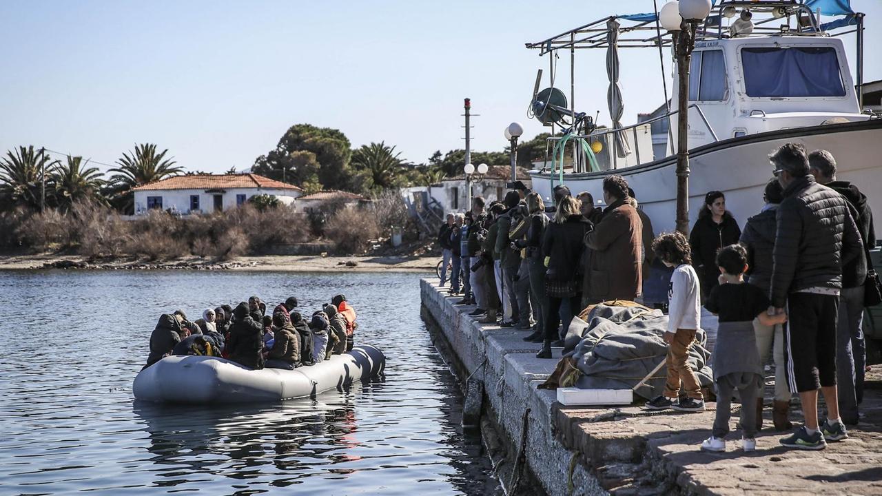 Ein Schlauchboot mit ca. 20 Menschen liegt vor einem Anlegepier, auf dem ähnlich viele Menschen aufgereiht stehen. Ein Schlauchboot mit ca. 20 Menschen liegt vor einem Anlegepier, auf dem ähnlich viele Menschen aufgereiht stehen.