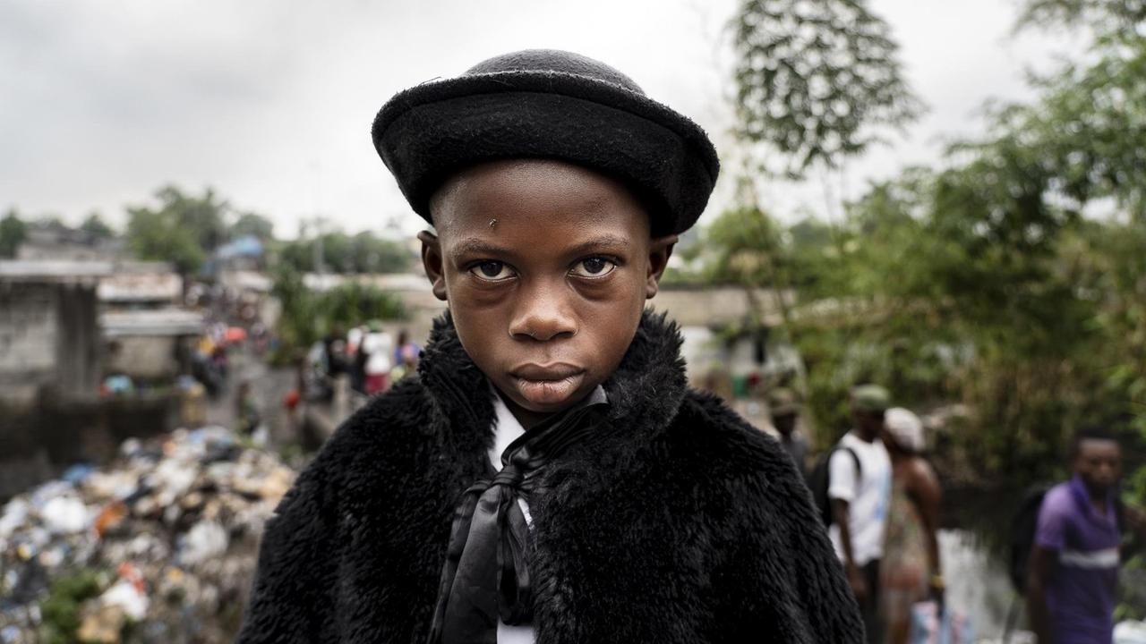 Ein kongolesischer Junge mit schwarzem Hut und Pelzmantel schaut direkt in die Kamera.