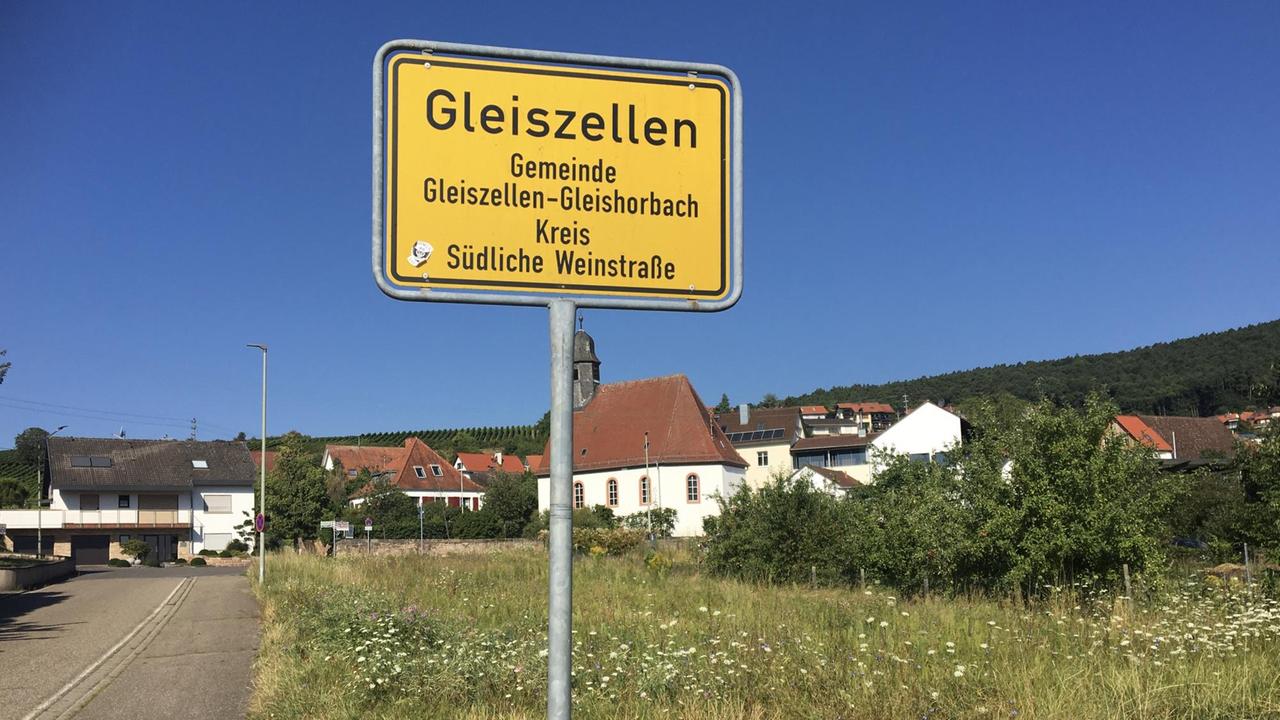 Das Bild zeigt ein gelbes Ortsschild an einer Dorfstraße vor sommerlich blauem Himmel. Das Bild zeigt ein gelbes Ortsschild an einer Dorfstraße vor sommerlich blauem Himmel.