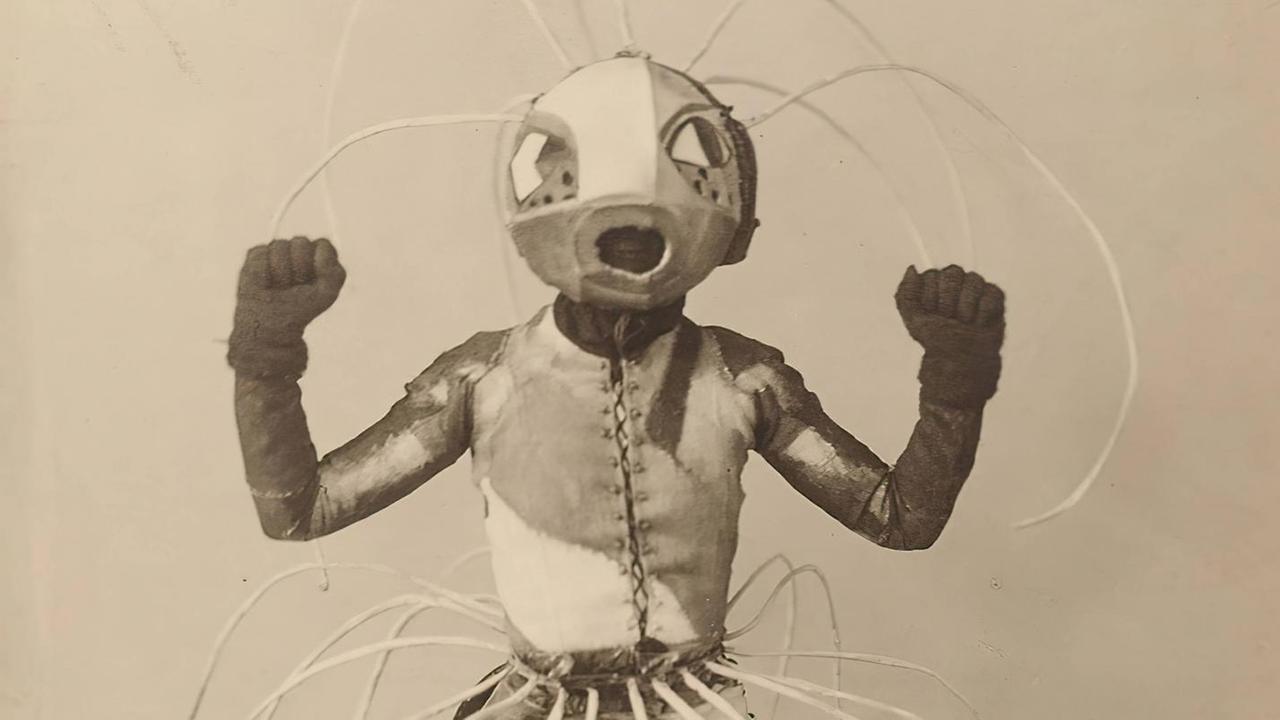 Ein Schwarzweiß-Foto zeigt eine breitbeinig stehende Roboter-ähnliche Puppe mit nach oben gereckten Armen