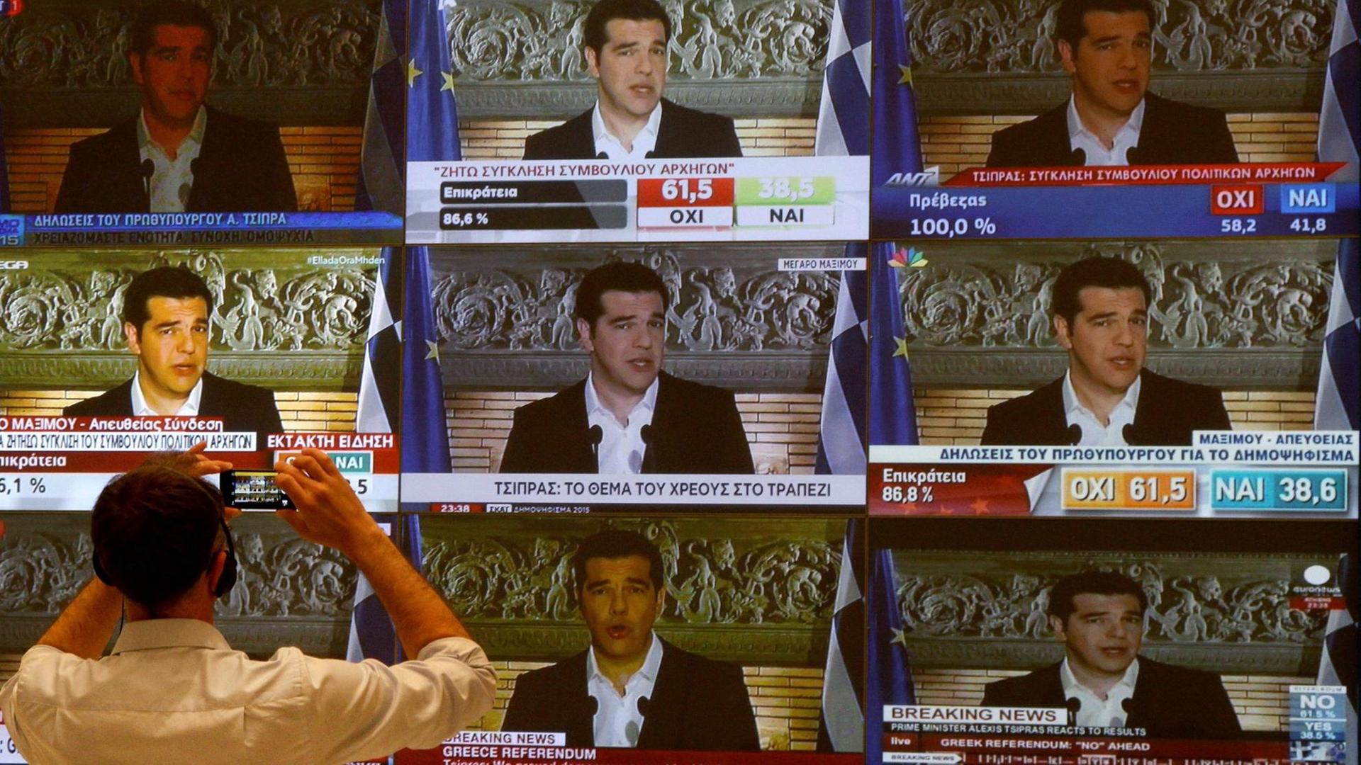 Ein Journalist steht vor insgesamt neun fernsehbildschirmen, auf denen Alexis Tsipras zu sehen ist. Ein Journalist steht vor insgesamt neun fernsehbildschirmen, auf denen Alexis Tsipras zu sehen ist.