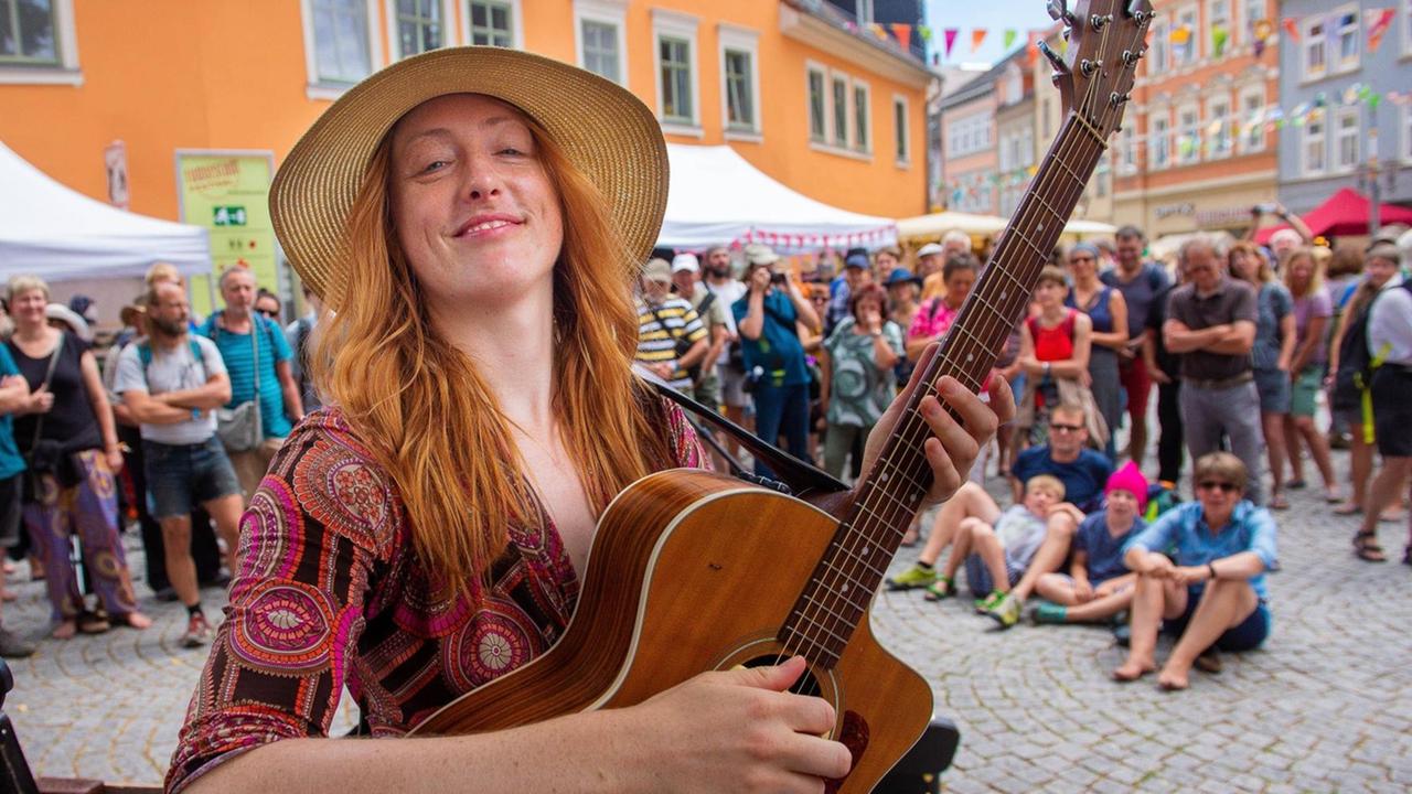 Eine Frau mit Gitarre steht auf einem Platz. Im Hintergrund sieht man das Publikum.