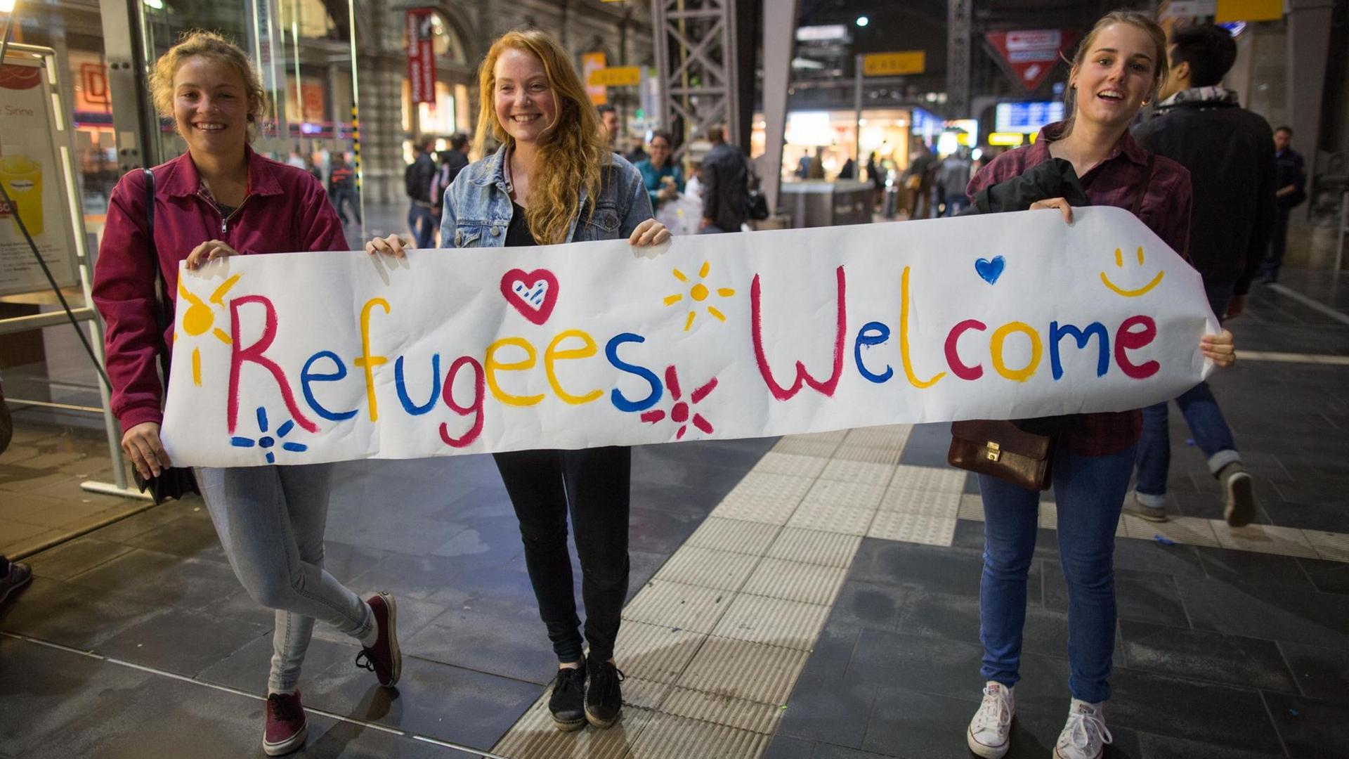 Drei junge Frauen stehen mit einem Begrüßungsplakat mit der Aufschrift "Refugees Welcome" für Flüchtlinge an den Gleisen. Drei junge Frauen stehen mit einem Begrüßungsplakat mit der Aufschrift "Refugees Welcome" für Flüchtlinge an den Gleisen.