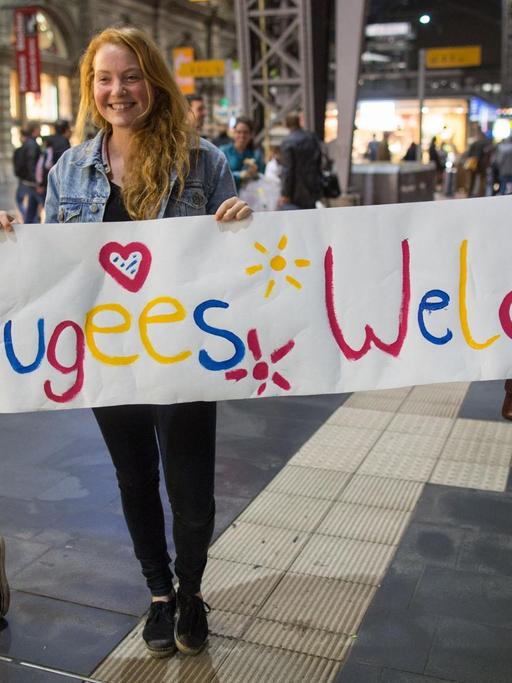 Drei junge Frauen stehen mit einem Begrüßungsplakat mit der Aufschrift "Refugees Welcome" für Flüchtlinge an den Gleisen.