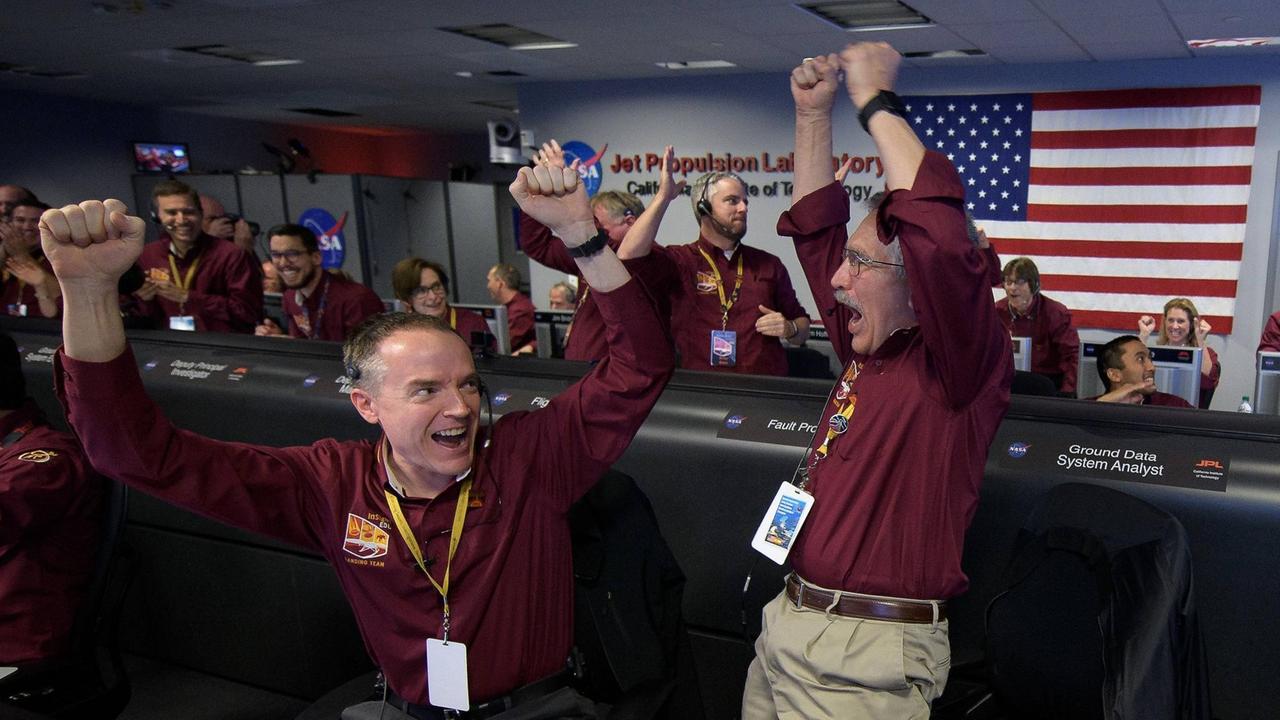 NASA-Mitarbeiter in weinroten Shirts jubeln und recken die Hände in die Höhe. NASA-Mitarbeiter in weinroten Shirts jubeln und recken die Hände in die Höhe.