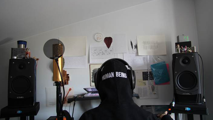Antye Greie im Heimstudio, sitzt mit dem Rücken zur Kamera, hat ein Kapuzen-Shirt mit der Aufschrift Human Being an und daürber Kopfhörer. Im Hintergrund sind zwei Lautsprecher zu sehen. 