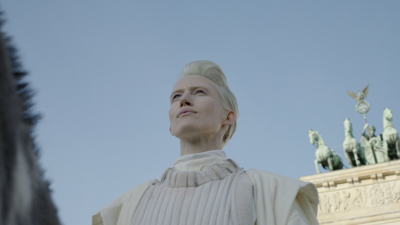 Filmstill aus "Malka Germania": Eine androgyne Figur vor dem Brandenburger Tor.