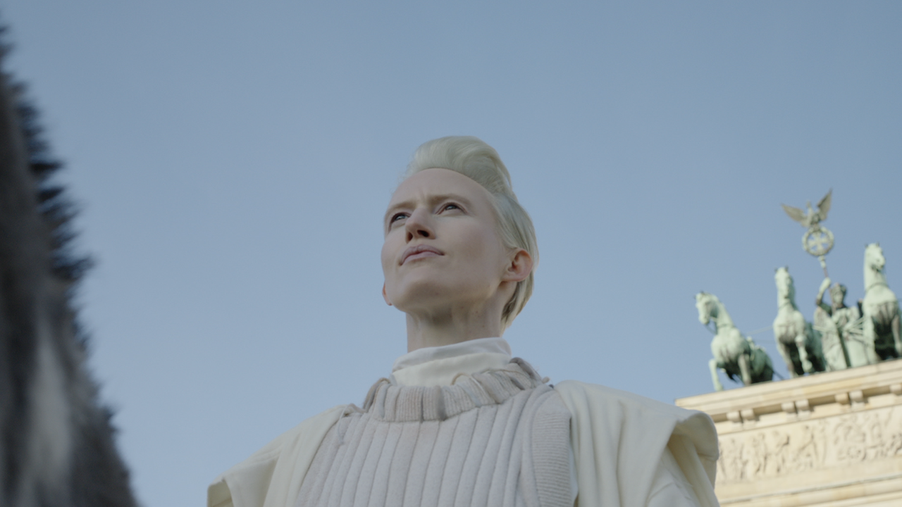 Filmstill aus "Malka Germania": Eine androgyne Figur vor dem Brandenburger Tor. Filmstill aus "Malka Germania": Eine androgyne Figur vor dem Brandenburger Tor.