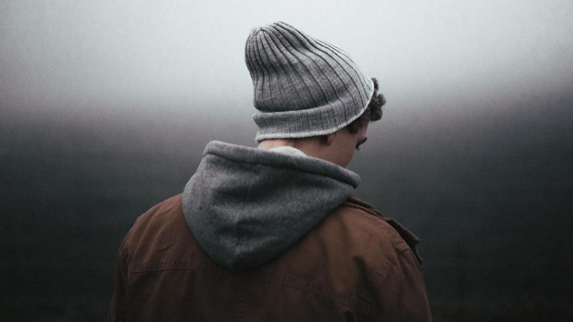 Ein junger Mensch mit Wollmütze, Kapuzenpulli und brauner Jacke schaut vor einem düsteren, nebligen Hintergrund zu Boden. Ein junger Mensch mit Wollmütze, Kapuzenpulli und brauner Jacke schaut vor einem düsteren, nebligen Hintergrund zu Boden.