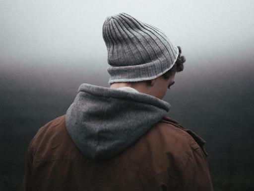 Ein junger Mensch mit Wollmütze, Kapuzenpulli und brauner Jacke schaut vor einem düsteren, nebligen Hintergrund zu Boden.