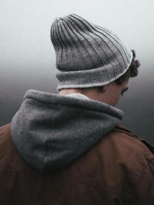 Ein junger Mensch mit Wollmütze, Kapuzenpulli und brauner Jacke schaut vor einem düsteren, nebligen Hintergrund zu Boden.
