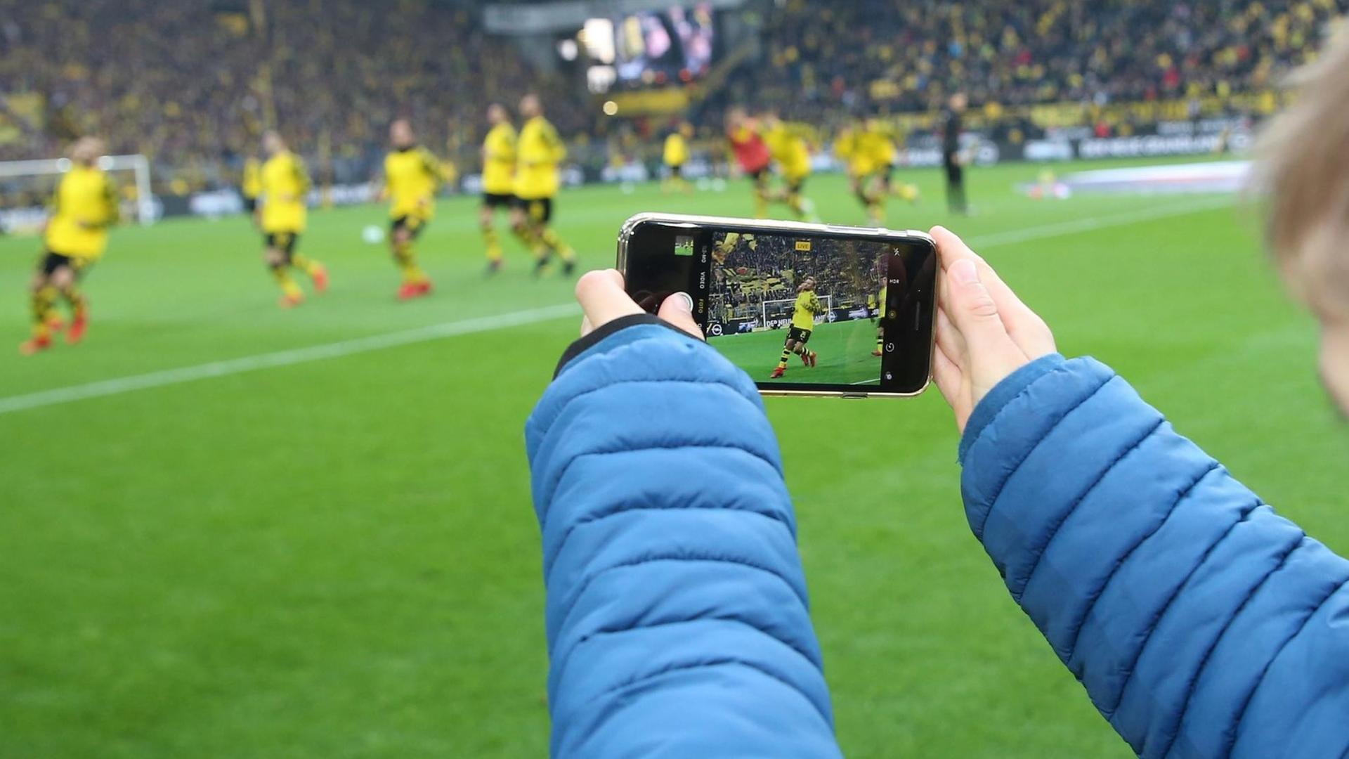 Ein junger Fußballfan filmt mit seinem Smartphone eine Szene eines Fußballspiels im Stadion