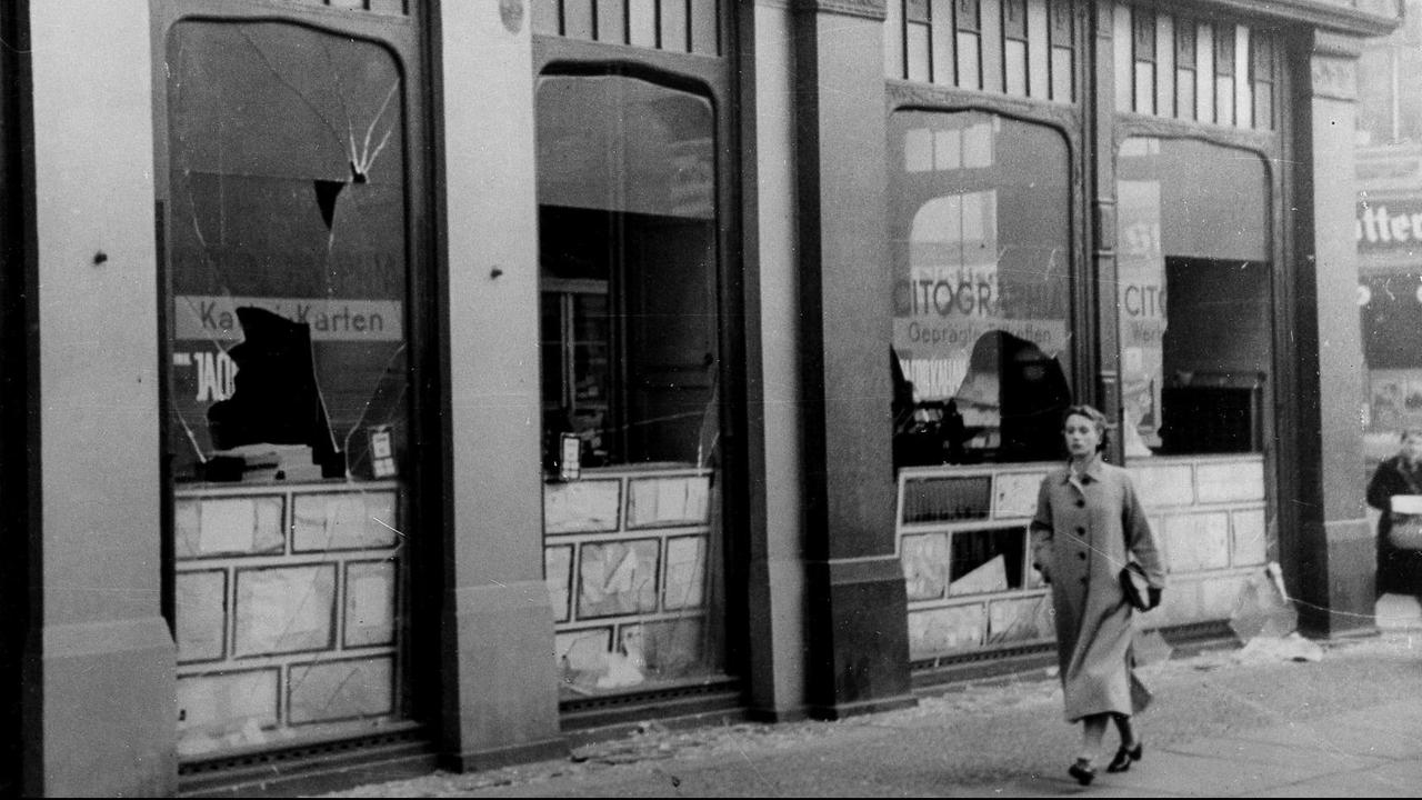 Eine Frau läuft in Berlin im Jahr 1938 an einem Geschäft vorbei, das von Juden geführt wurde. Die Scheiben sind zerbrochen.