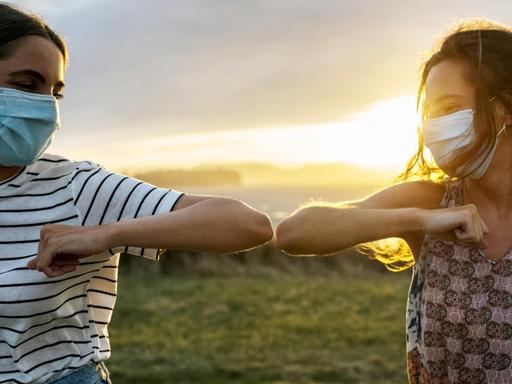 Zwei junge Mädchen mit Gesichtsmaske begrüssen sich mit einem Ellenbogengruss. Zwei junge Mädchen mit Gesichtsmaske begrüssen sich mit einem Ellenbogengruss.