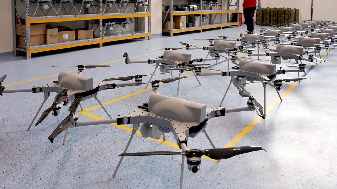 Aufgereihte Drohnen stehen nebeneinander auf einem blauen Linoliumboden in einer Lagerhalle.