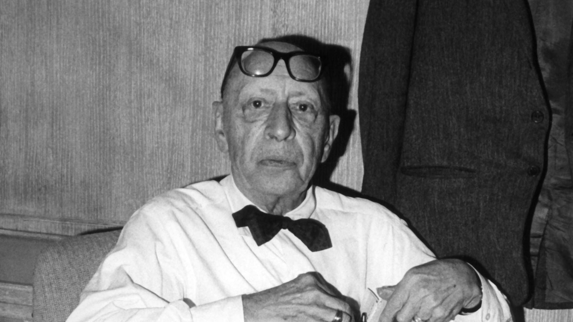 Der Komponist Igor Strawinsky mit weißem Hemd, schwarzer Fliege und einer Brille auf der Stirn.