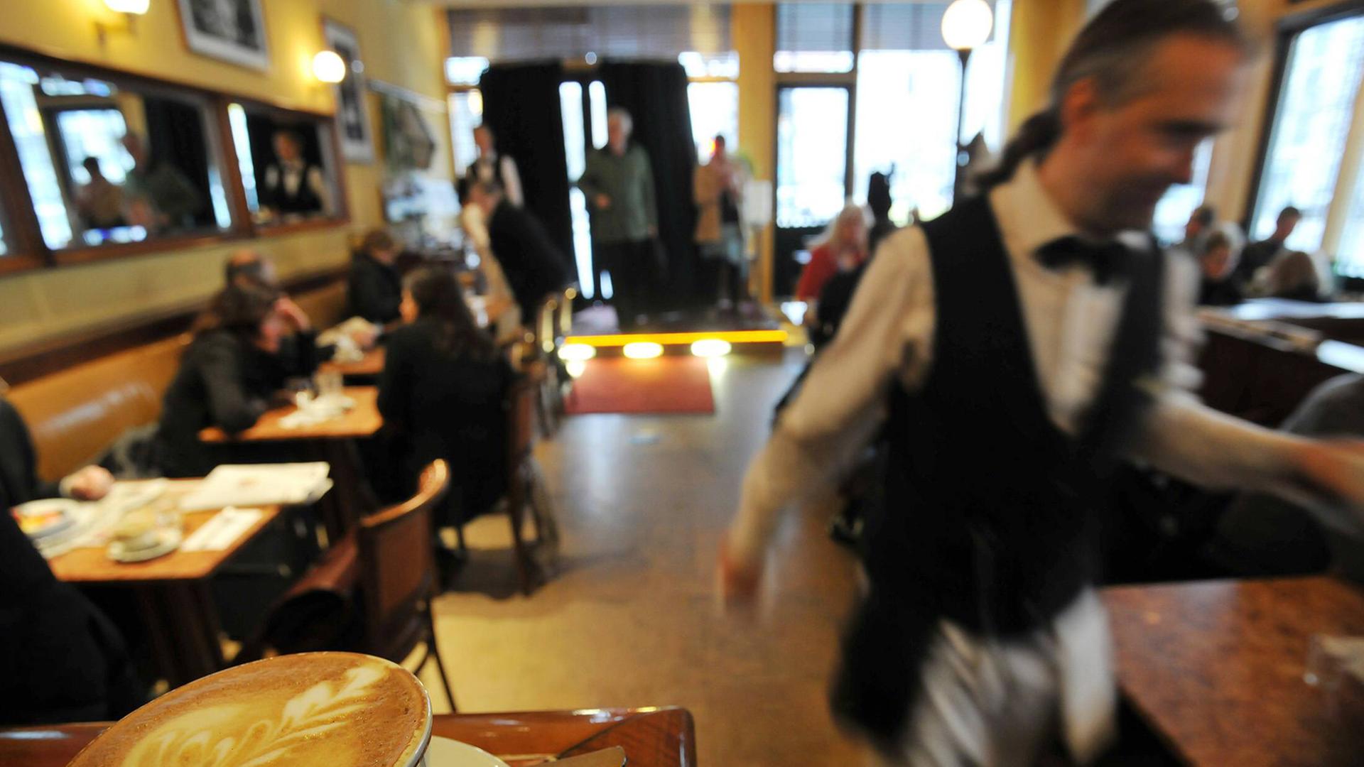 Blick in ein Restaurant, auf einer Theke im Vordergrund steht ein Kaffee, ein Kellner läuft durchs Bild.