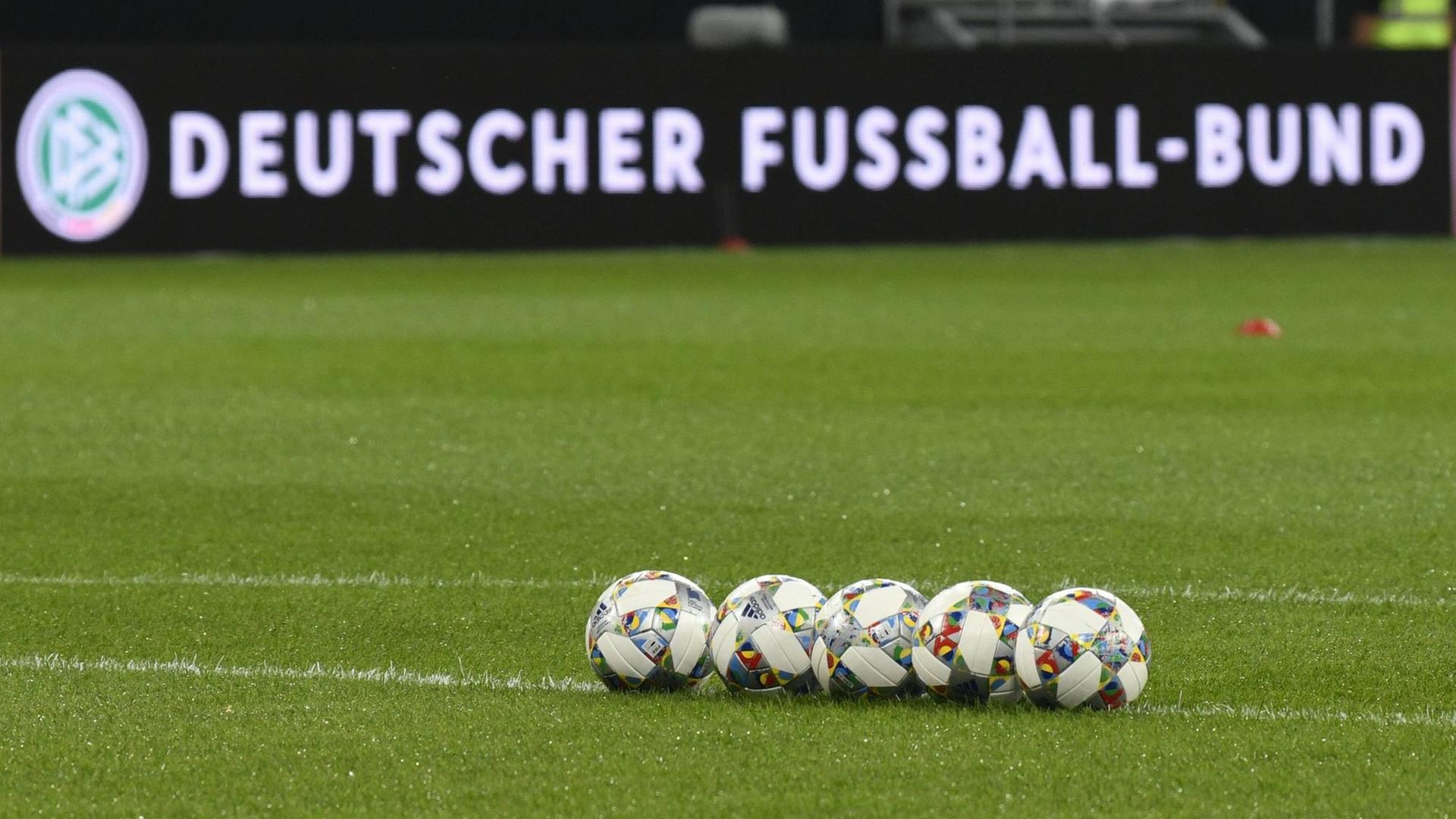 Mehrere Fussbälle liegen aufgereiht auf dem Rasen, im Hintergrund eine Bandenwerbung mit Logo und Schriftzug "Deutscher Fussball-Bund"