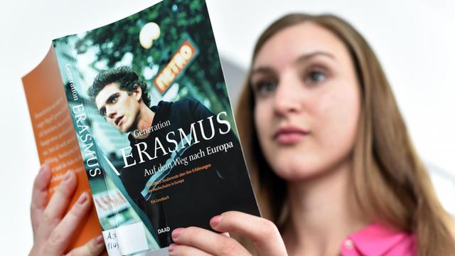 Eine Studentin liest in einem Buch über das Erasmus-Programm. Man sieht den Buch-Umschlag im Vordergrund, das Gesicht der jungen Frau unscharf im Hintergrund.