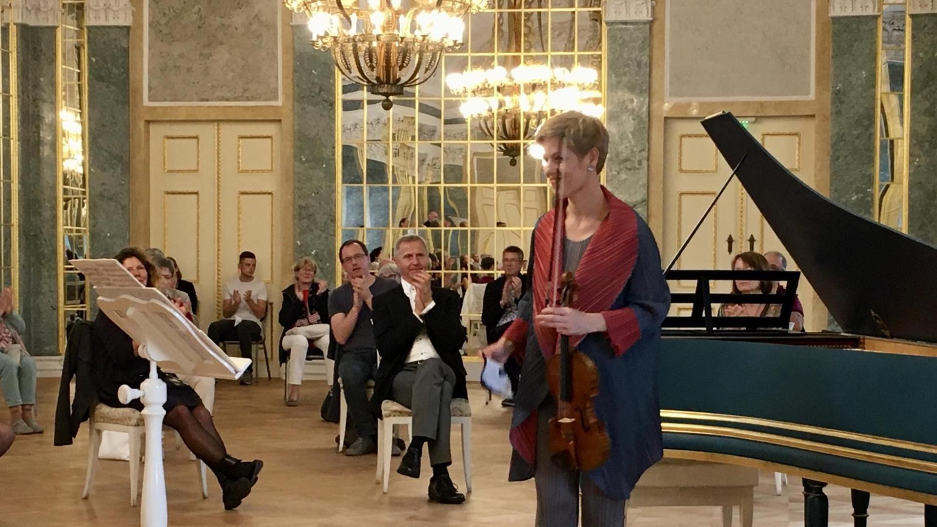 Eine Frau mit kurzen, blonden Haaren hält eine Geige in der linken Hand und steht in einem Schlosssaal vor einem Cembalo. Um sie herum verteilt sitzen Menschen mit viel Abstand und applaudieren. Eine Frau mit kurzen, blonden Haaren hält eine Geige in der linken Hand und steht in einem Schlosssaal vor einem Cembalo. Um sie herum verteilt sitzen Menschen mit viel Abstand und applaudieren.