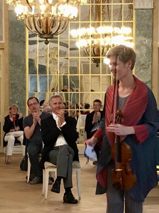Eine Frau mit kurzen, blonden Haaren hält eine Geige in der linken Hand und steht in einem Schlosssaal vor einem Cembalo. Um sie herum verteilt sitzen Menschen mit viel Abstand und applaudieren.