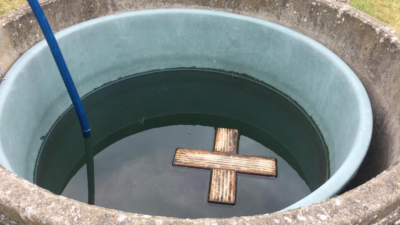 In einem mit Wasser gefüllten Bottich schwimmt ein kleines Kreuz aus Holz an der Wasseroberfläche. In einem mit Wasser gefüllten Bottich schwimmt ein kleines Kreuz aus Holz an der Wasseroberfläche.