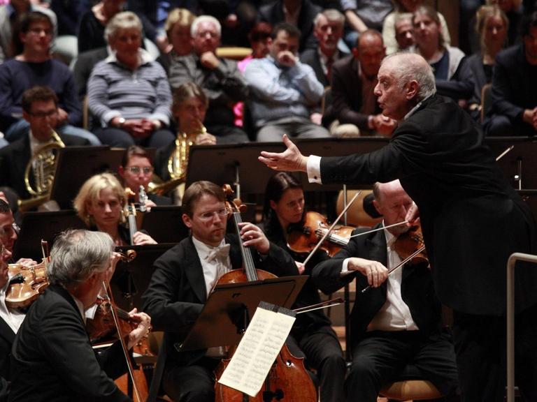 Daniel Barenboim mit einer ausladenden Geste vor dem Orchester, im Hinergrund ist Publikum zu sehen.